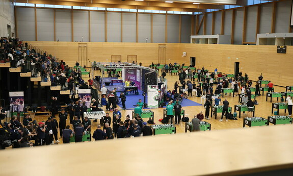 Halle mit mehreren Menschen die am Tischkicker ihr Glück versuchen. Auf der linken Seite tobende Menschen.